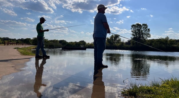 Two men fishing along the water’s edge beneath a partly cloudy sky, with their reflections mirrored in the calm water.